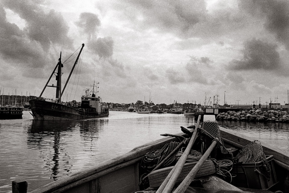 black and white photograph taken from inside a small rowboat on a harbor, under a cloudy sky