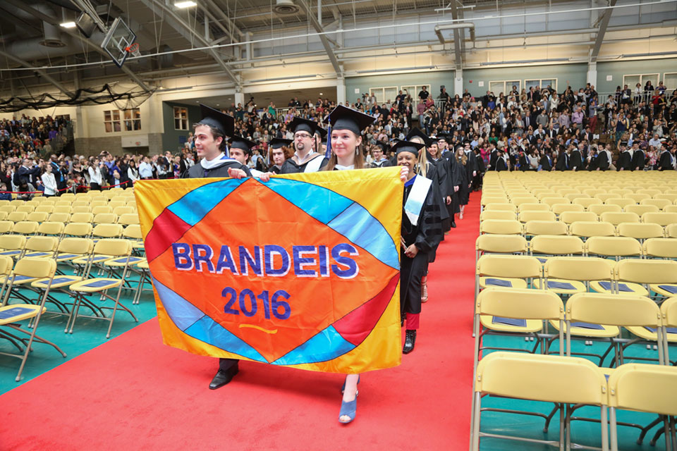 Students in caps and gowns walk into Gosman with a Brandeis 2016 banner