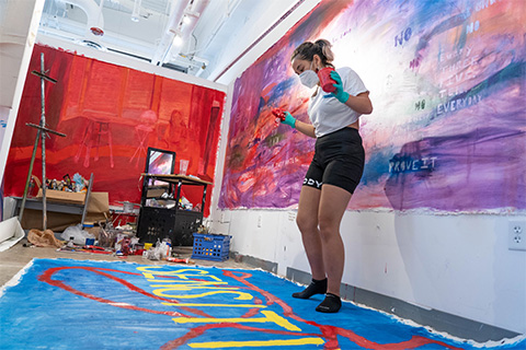 Bucu standing on top and looking down at artwork that has yellow letters, red lines, and a blue background; the walls in the background are vibrant with large paintings on them