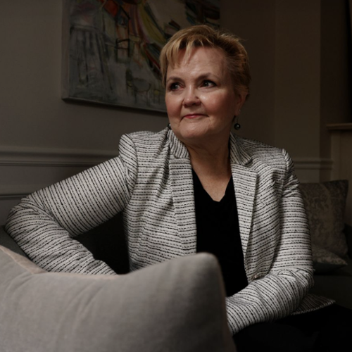 Barbara Clarke smiling and looking into the distance while sitting on a couch and holding a pillow.