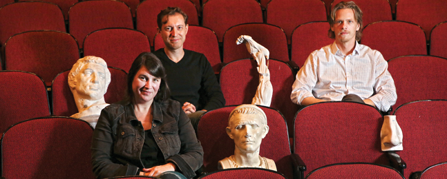 Cameron Anderson, Dmitry Troyanovsky and Joel Christensen sitting in red theater seats surrounded by bits of marble sculpture of human figures.