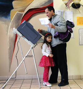 Photo of a little girl standing next to her father as he casts his ballot at a voting machine.