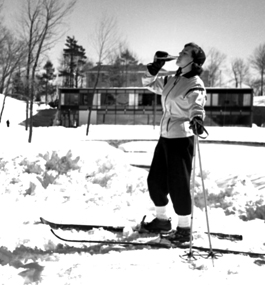 A female skier on campus in 1956, drinking from a bottle against the backdrop of a snow-covered campus.