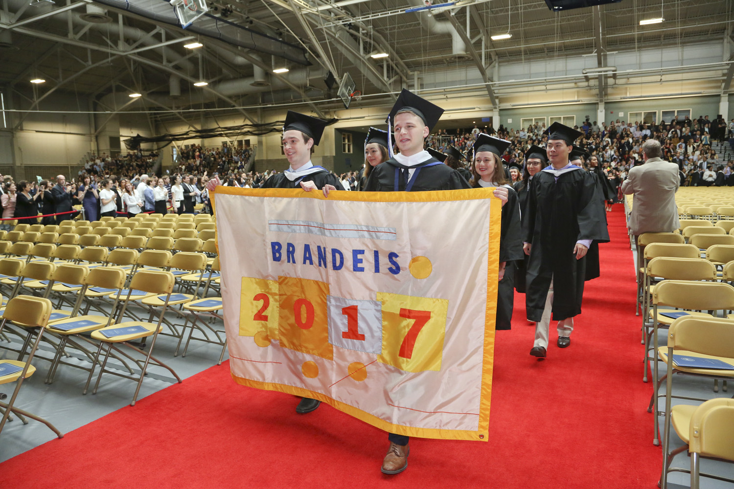 students in caps and gowns, holding a Brandeis 2017 banner, marching into Gosman Center for Commencement