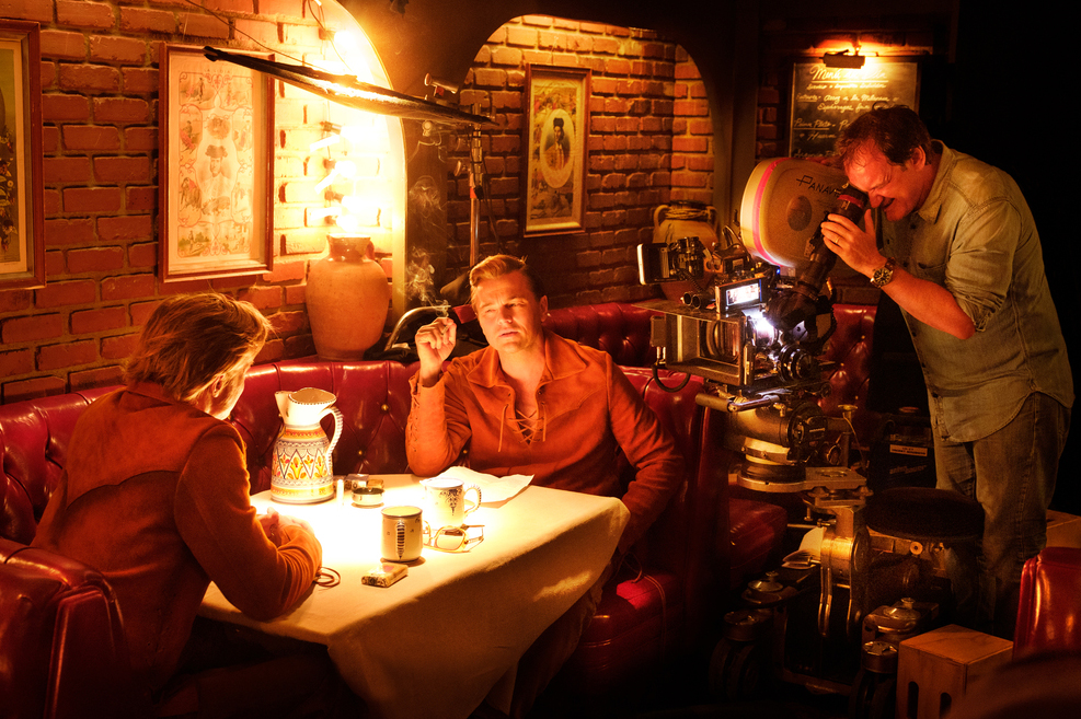 Film director Quentin Tarantino on a set filming Brad Pitt (back to camera) and Leonardo DiCaprio; they are sitting at a table in a red restaurant booth