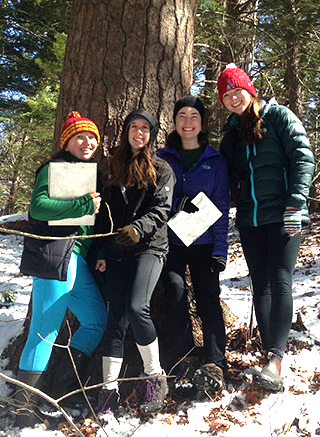 Four women students in winter coats and hats in front of a hemlock tree