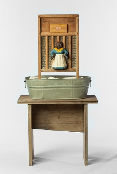 sculpture using old scrub board, fronted by an old-style stereotypical representation of African-American woman in apron and handkerchief holding pistols, stood in an aluminum washtub on wooden table, titled "Supreme Quality" by artist Betye Saar