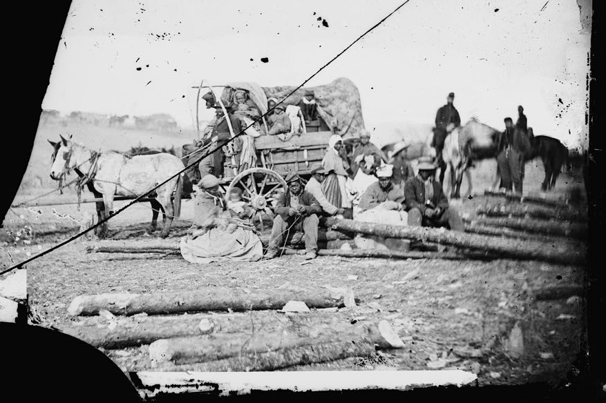 a damaged black and white photo of a group of escaped enslaved people around a covered wagon pulled by horses with sociers in the background