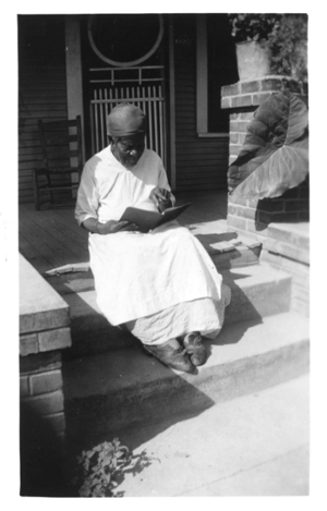 Black and white photo of Mary Armstrong, former slave, sitting on a porch stoop in 1937 with a book in her hand