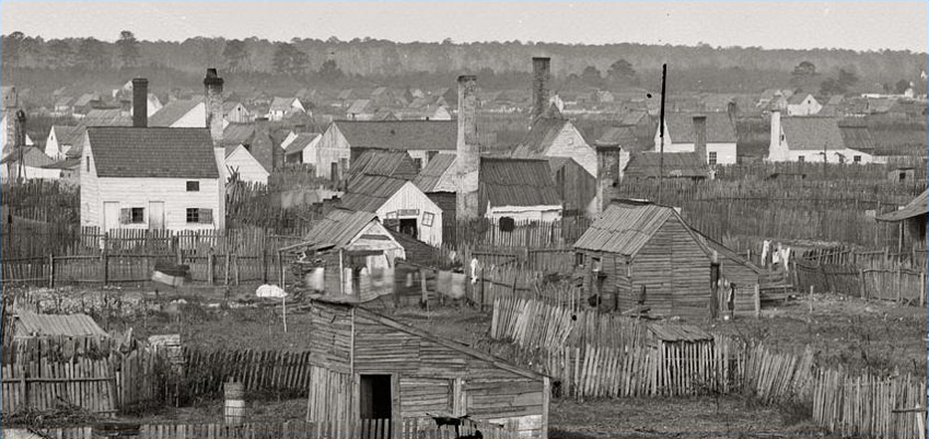 black and white photo of Slabtown, part of a refugee camp for escaped enslaved people during the Civil War. Some painted houses with chimneys and other shacks made of wood divided by slab fences 