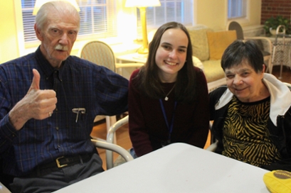 an older man in a blue shirt gives a camera a thumbs up, with his arm around a female student and his wife, an older woman, on the right. All are seated at a table.
