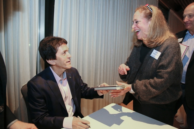Mitch Albom '79 in a jacket seated at a table, hands a book to a smiling woman with long hair.