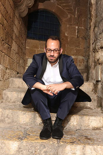 composer Yair Klartag in a suit with open shirt collar sits on a step in a stone stairway