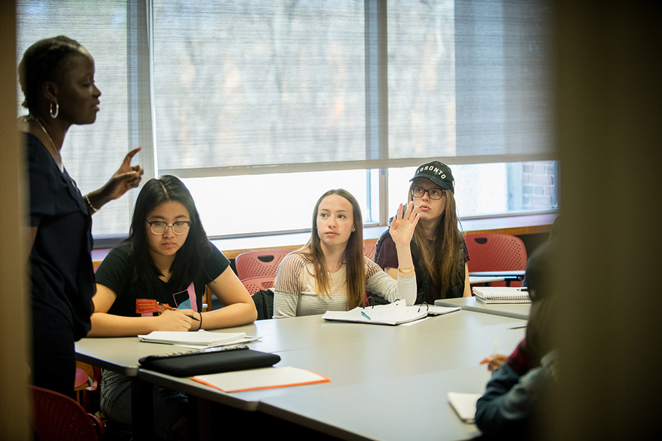 A female professor speaks to several female students around a table, with one raising her hand.