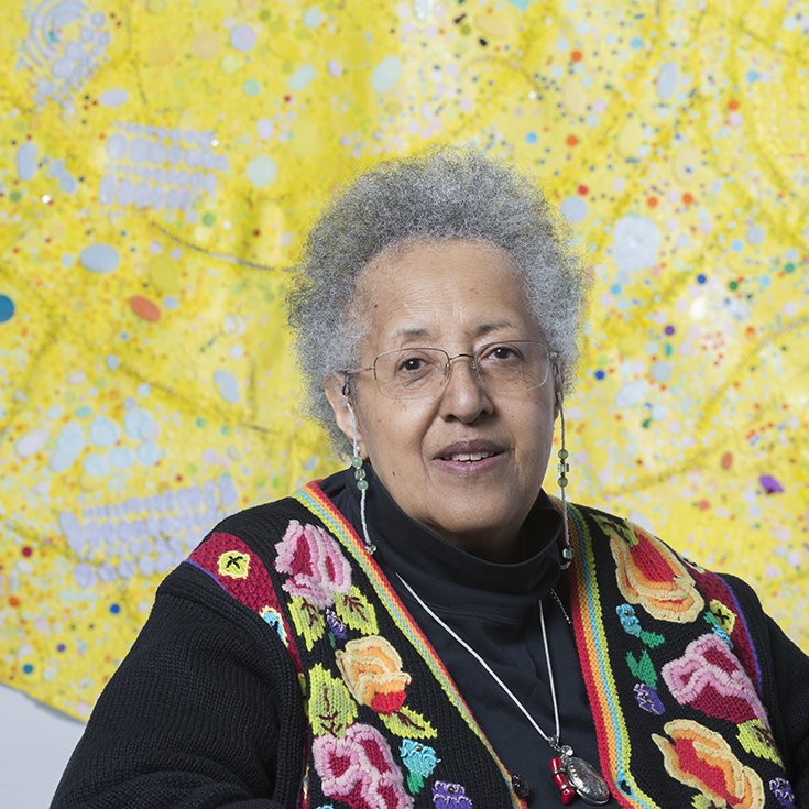 Head shot of Howardana Pindell in front of one of her paintings, a bright yellow abstraction