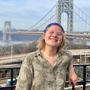 picture of a young smiling blonde person with a long New York bridge in the background