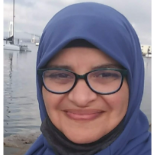 A headshot of a smiling woman with a blue hijab and black glasses, with water in the background