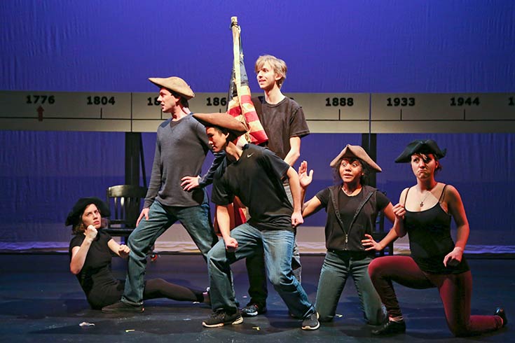 Group of students on stage, some kneeling and some standing, with an American Flag in the center.