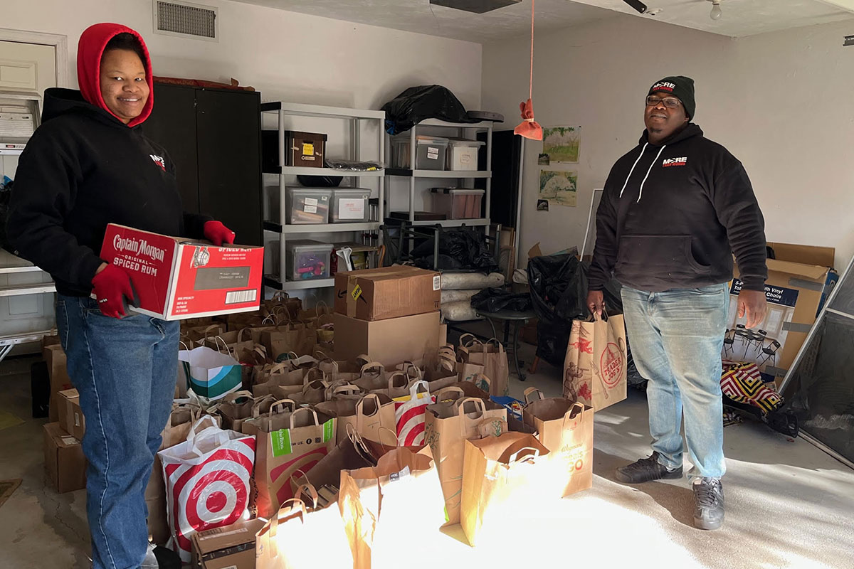 Two volunteers stand smiling in a garage surrounded by paper bags filled with books