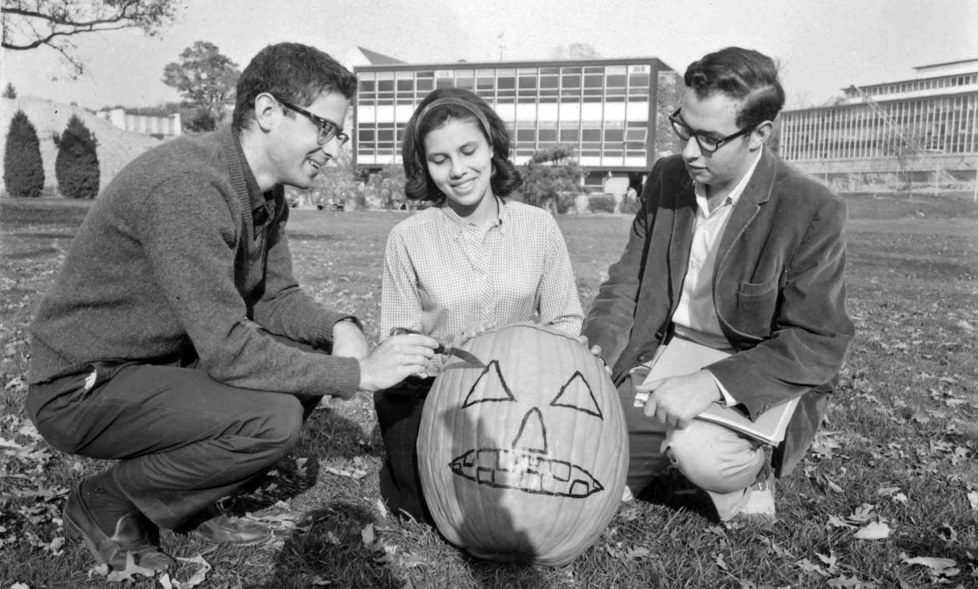 Students sit around a carved pumpkin sitting on the grass