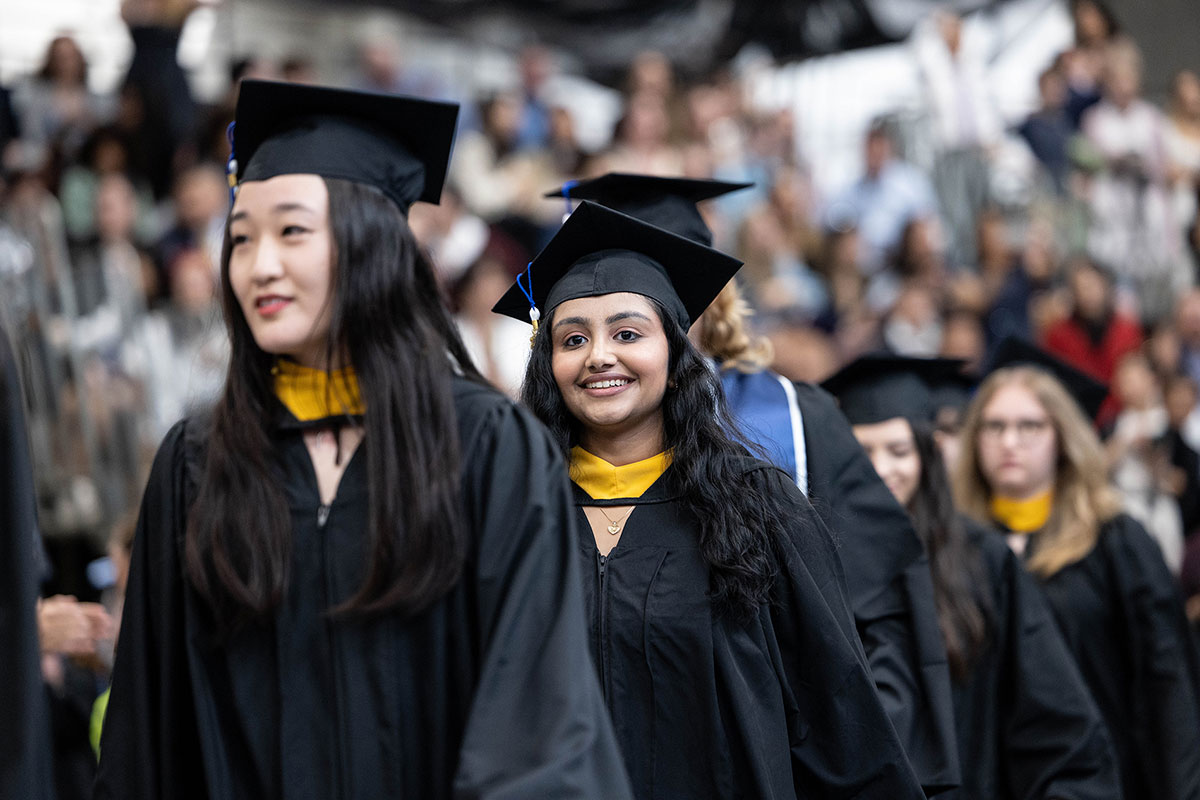 Student dressed in caps and gowns smile and walk in a graduation ceremony.