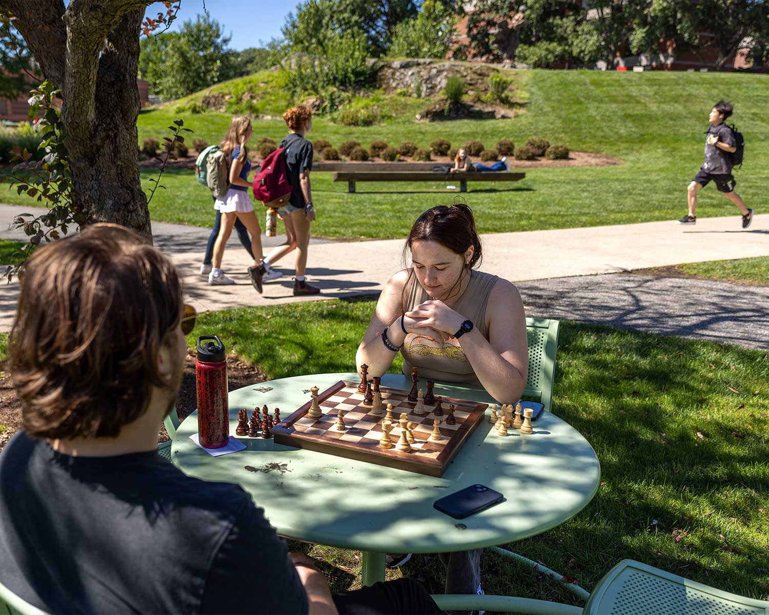 Two students play chess on a table beneath a tree.