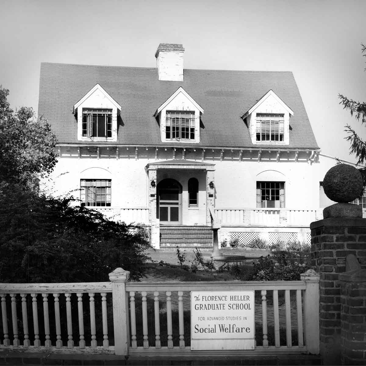 A black and white photo of a white house. Sign on a fence reads: The Florence Heller Graduate School for Advanced Studies Social Welfare