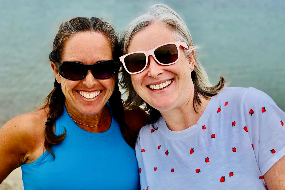 Two women wearing sunglasses and smiling with the ocean in the background. 