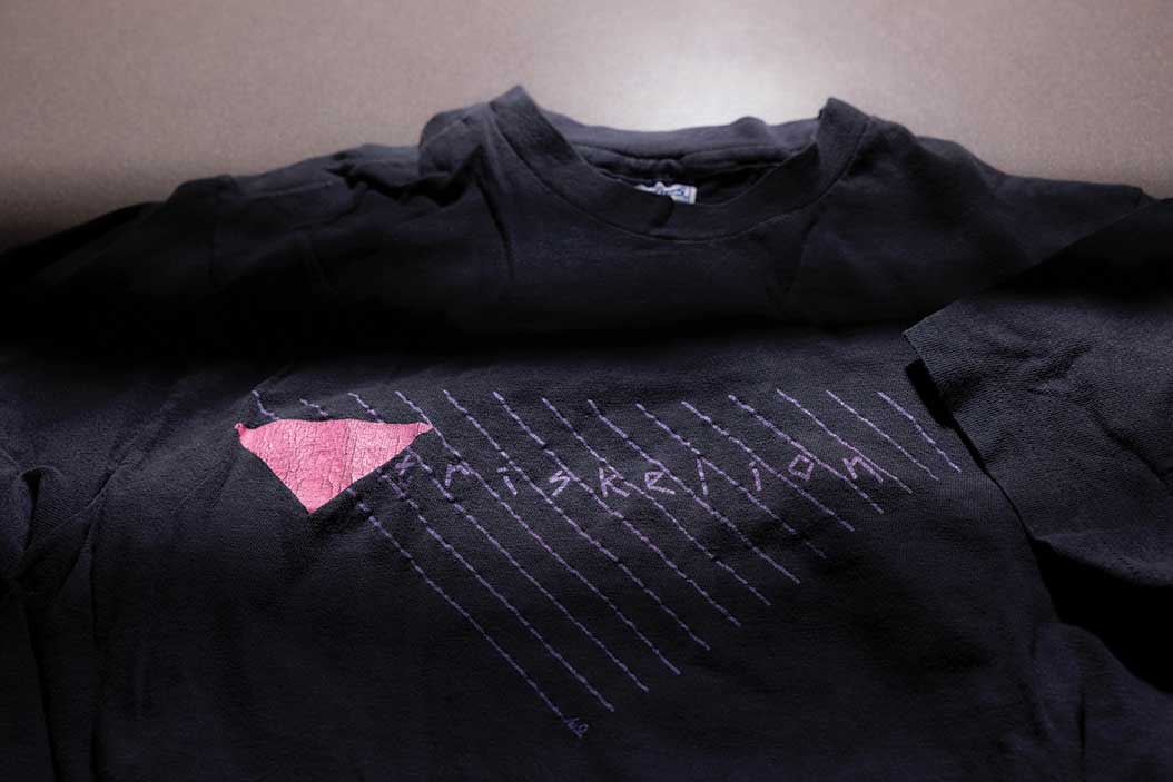 A shadowy photo of a dark T shirt emblazoned with a pink triangle and the name "Triskelion."