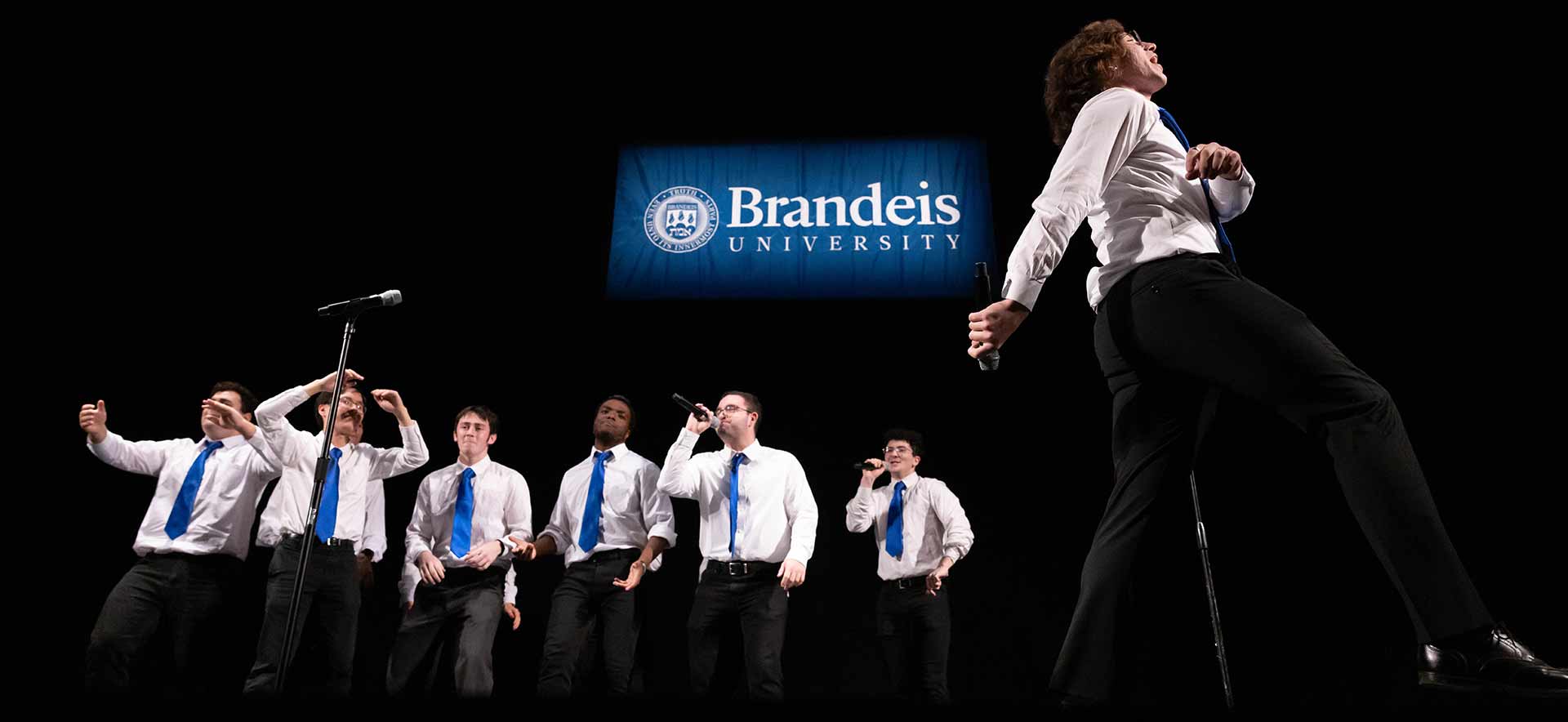People perform on stage wearing dark pants, white collared shirts and blue ties. A Brandeis banner hangs in the background.