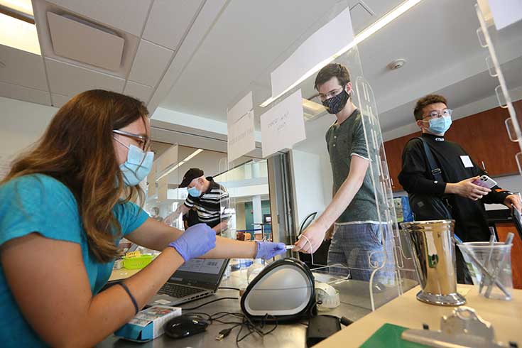 A person in a mask hands a COVID test to a person wearing a mask behind plexiglass