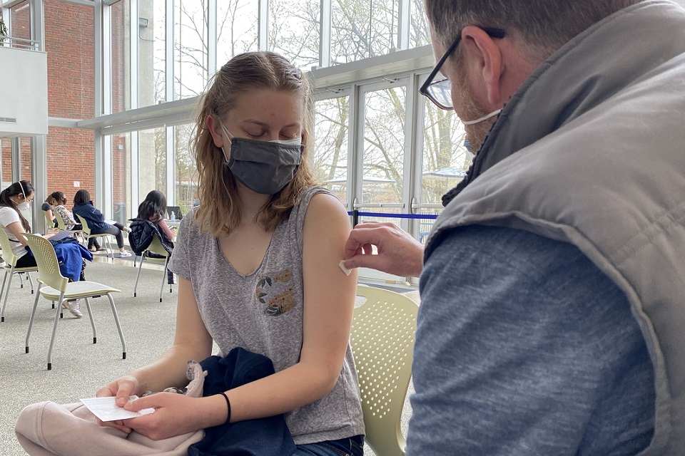 Woman, masked and seated with man wiping an alcohold swab on her arm prior to giving her a vaccine.