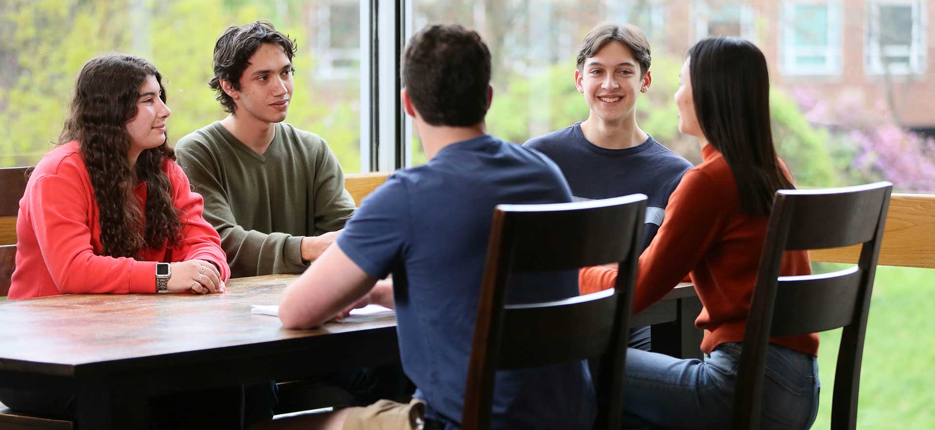 Five students seated at a table in conversation, the Brandeis campus is seen in the background