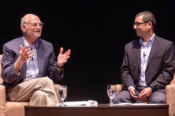 Michael Rosbash speaking while seated next to Vipin Suri on a stage