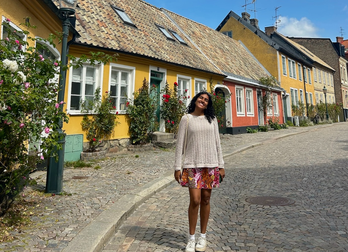 Anina stands on a cobblestone street in front of a bright yellow house on a sunny day in Copenhagen.