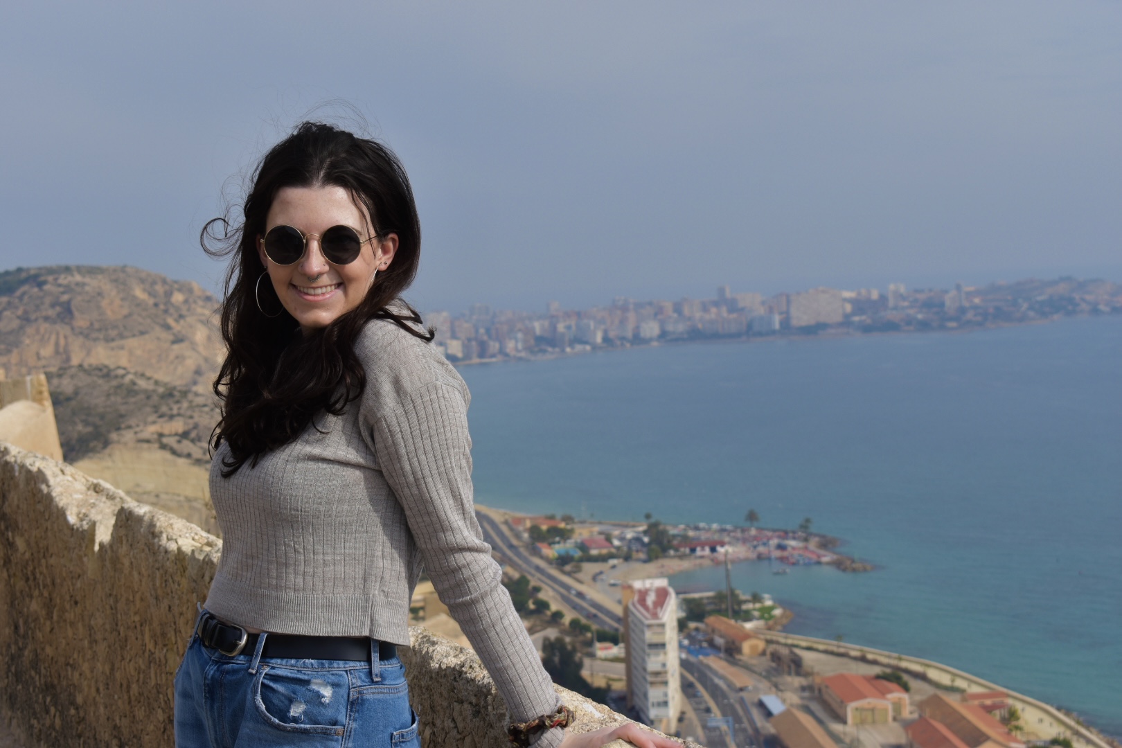Aileen smiling in sunglasses standing against a stone cliffside with the water beyond.