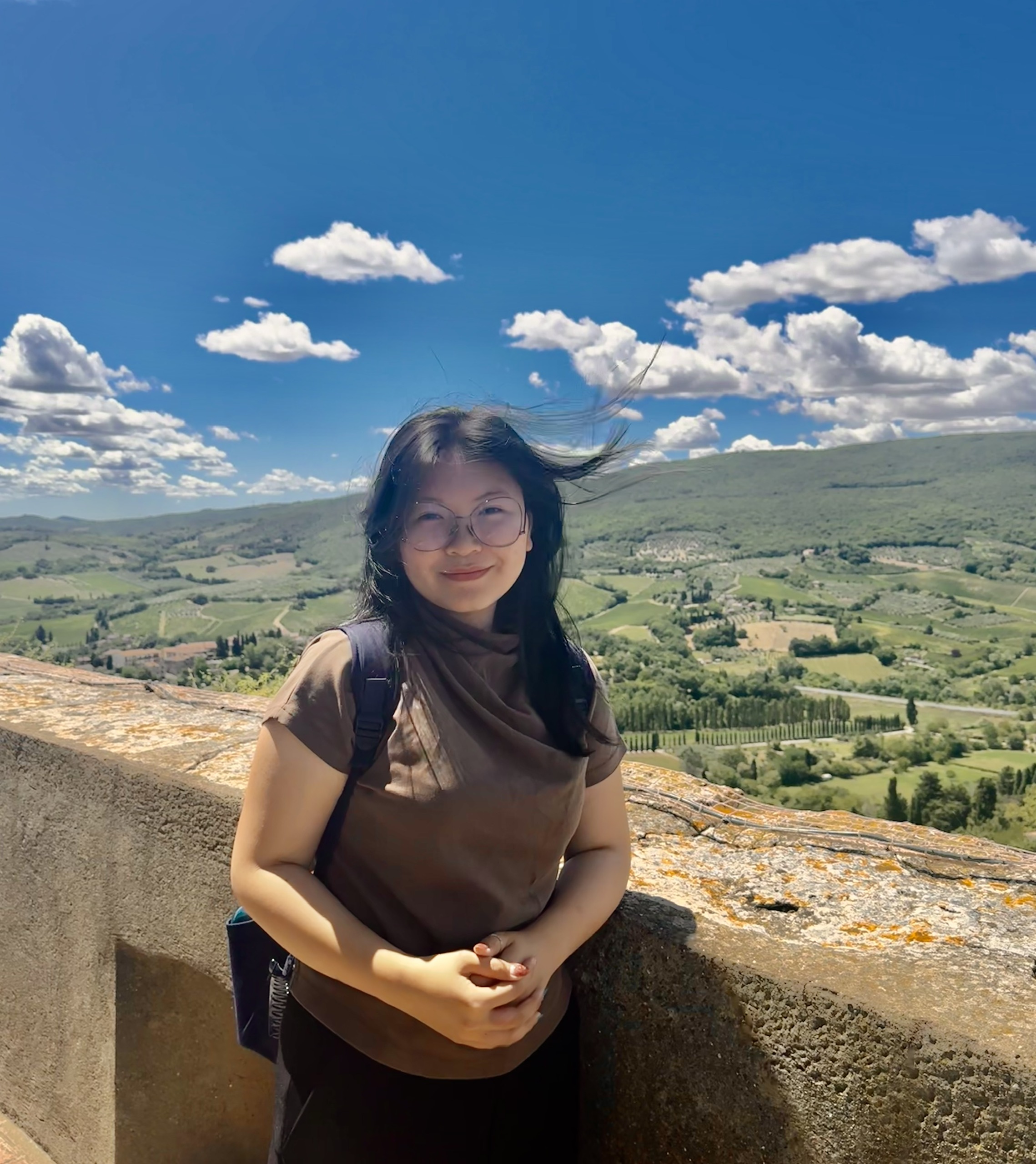 Cate stands against a stone balcony smiling on a sunny day in Italy, with green rolling hills behind her. Her hair flutters in the breeze.