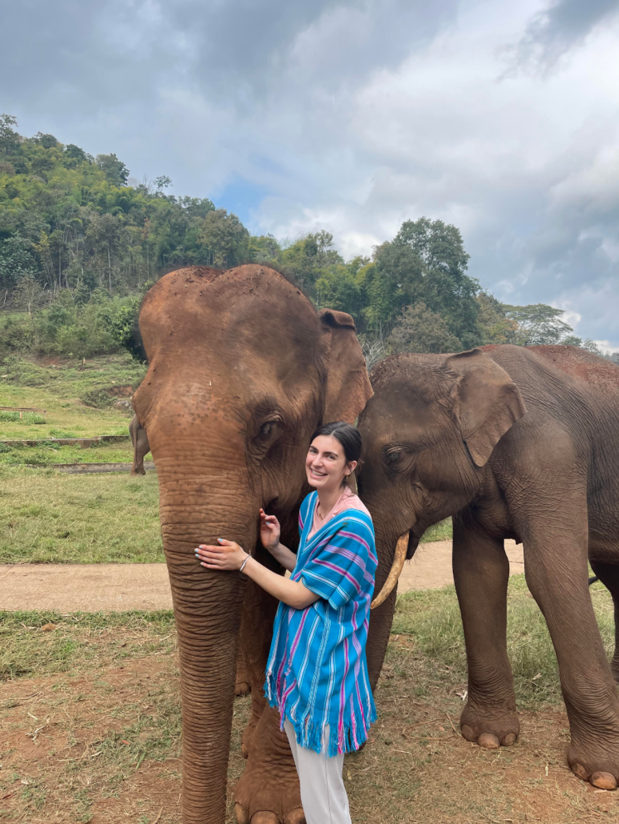 Ivy smiles while standing next to an elephant, gently holding its tusk.