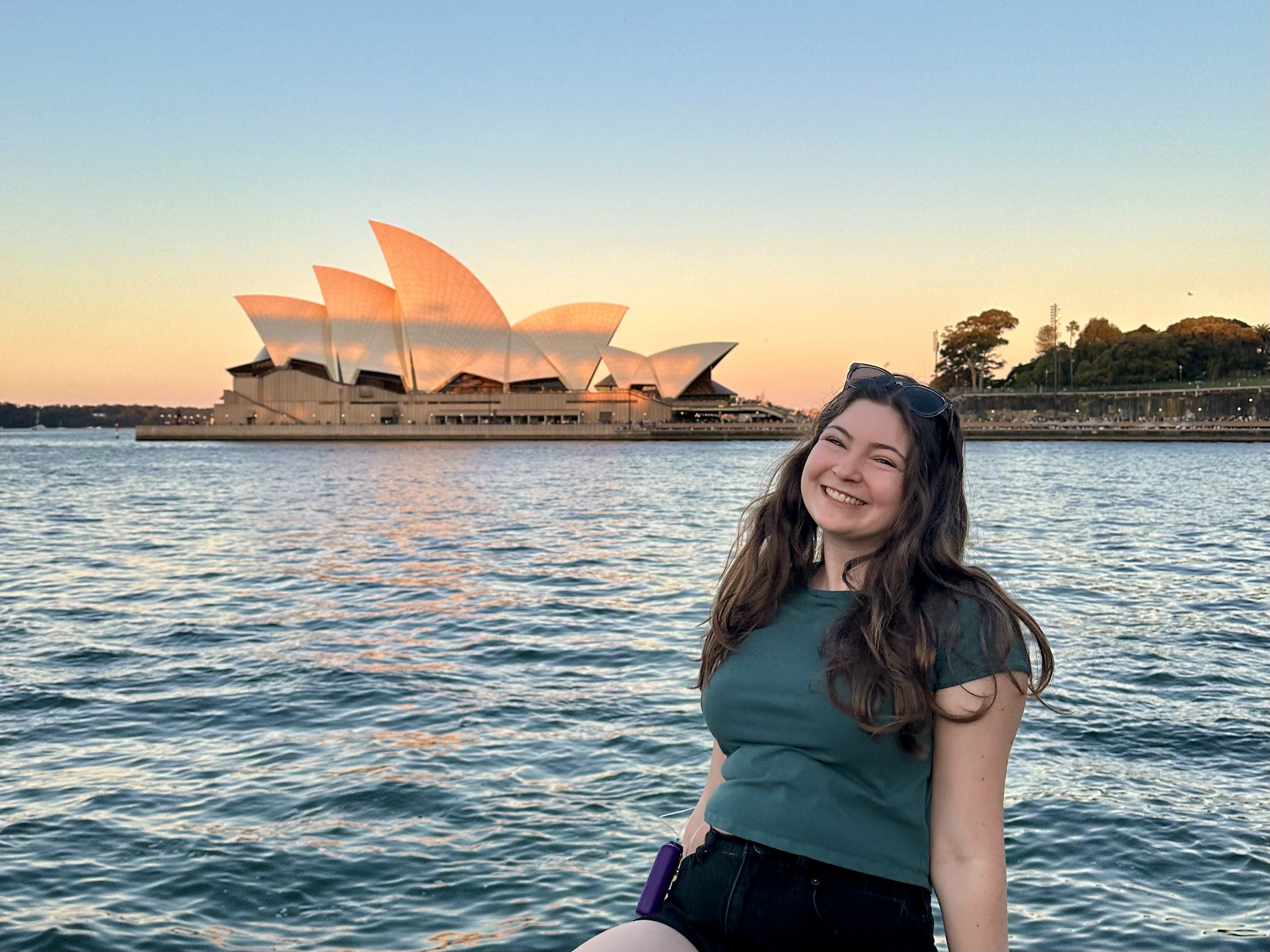 Ella smiles in the foreground with the Sydney Opera House and water at dusk in the background.