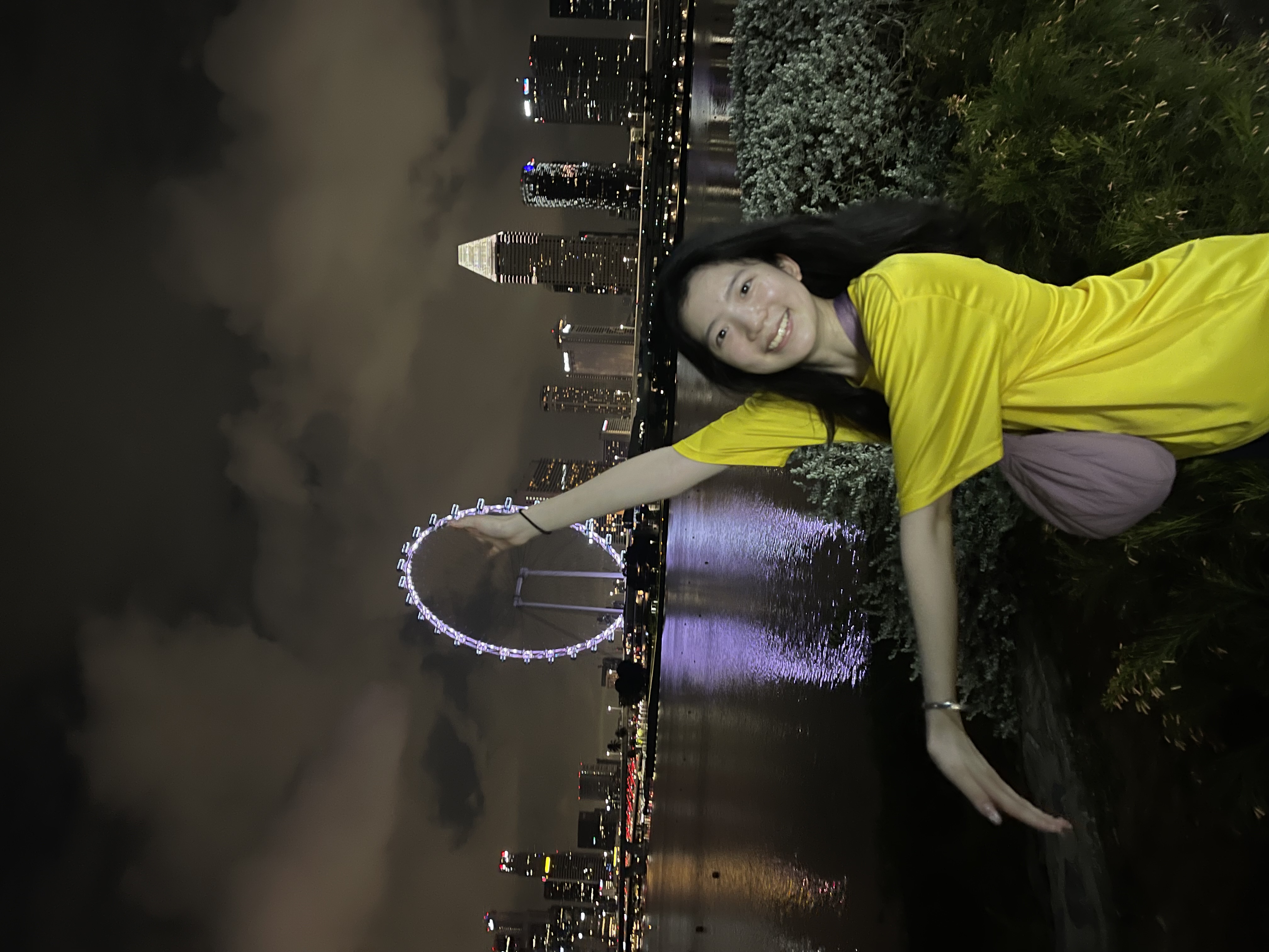 Emily wears a bright yellow shirt and gestures toward a lit-up ferris wheel on a night in Singapore.