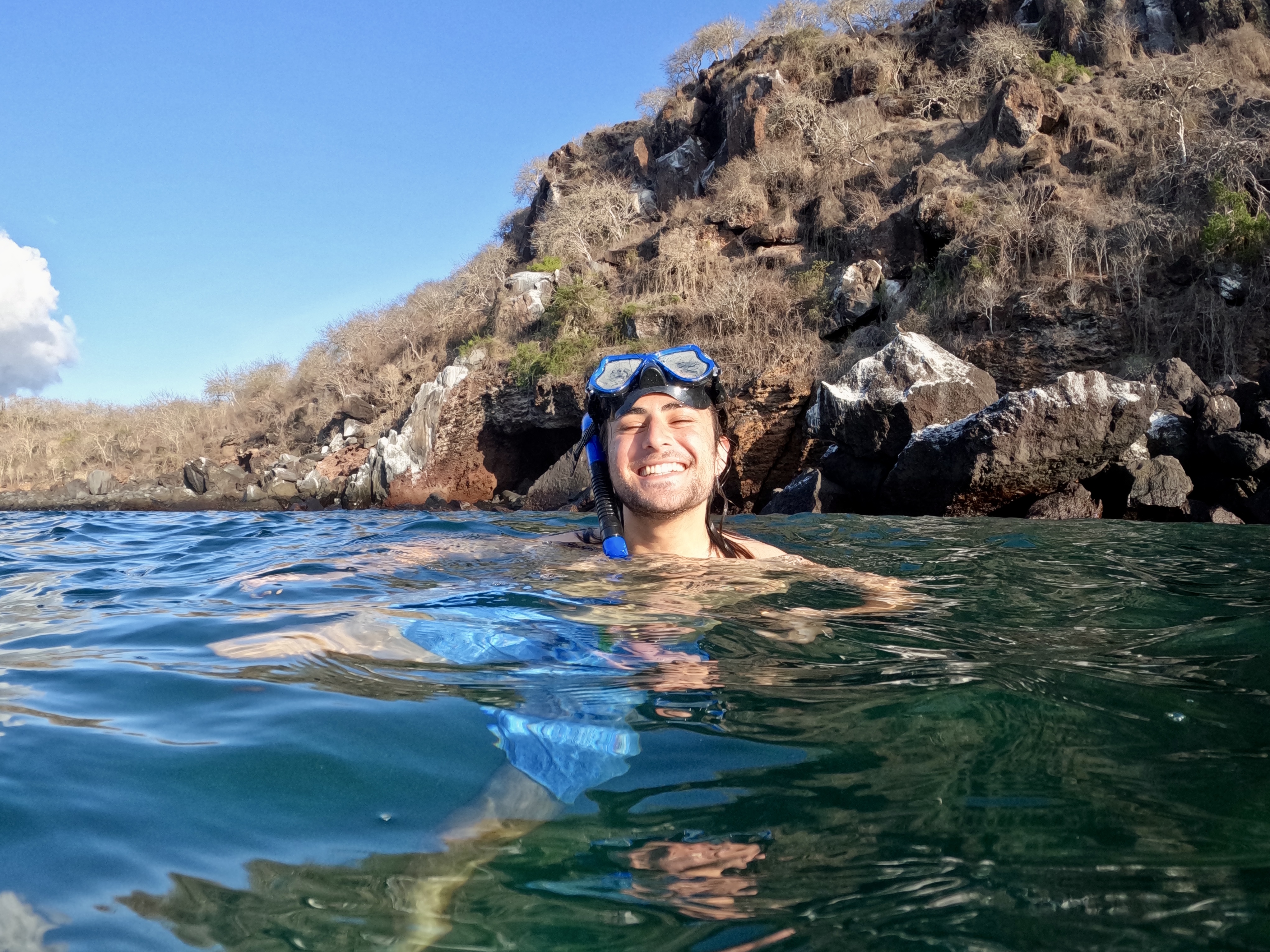 Saul grins as his head peeks out of the clear blue water, with the cliffside behind him. Goggles are perched on his head.