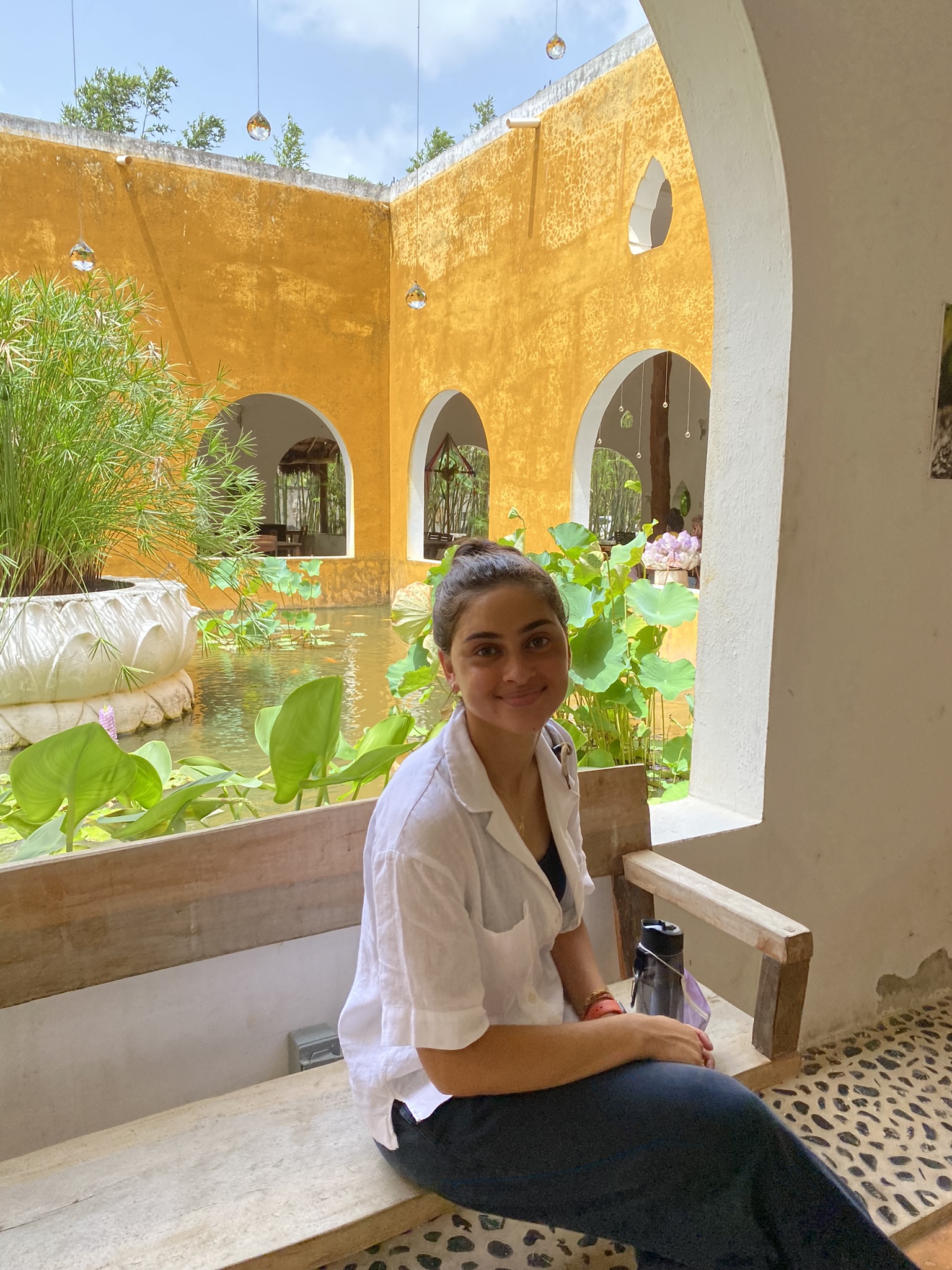 Yahni sits on a bench in front of a sunny courtyard with lush green plants and bright yellow walls.