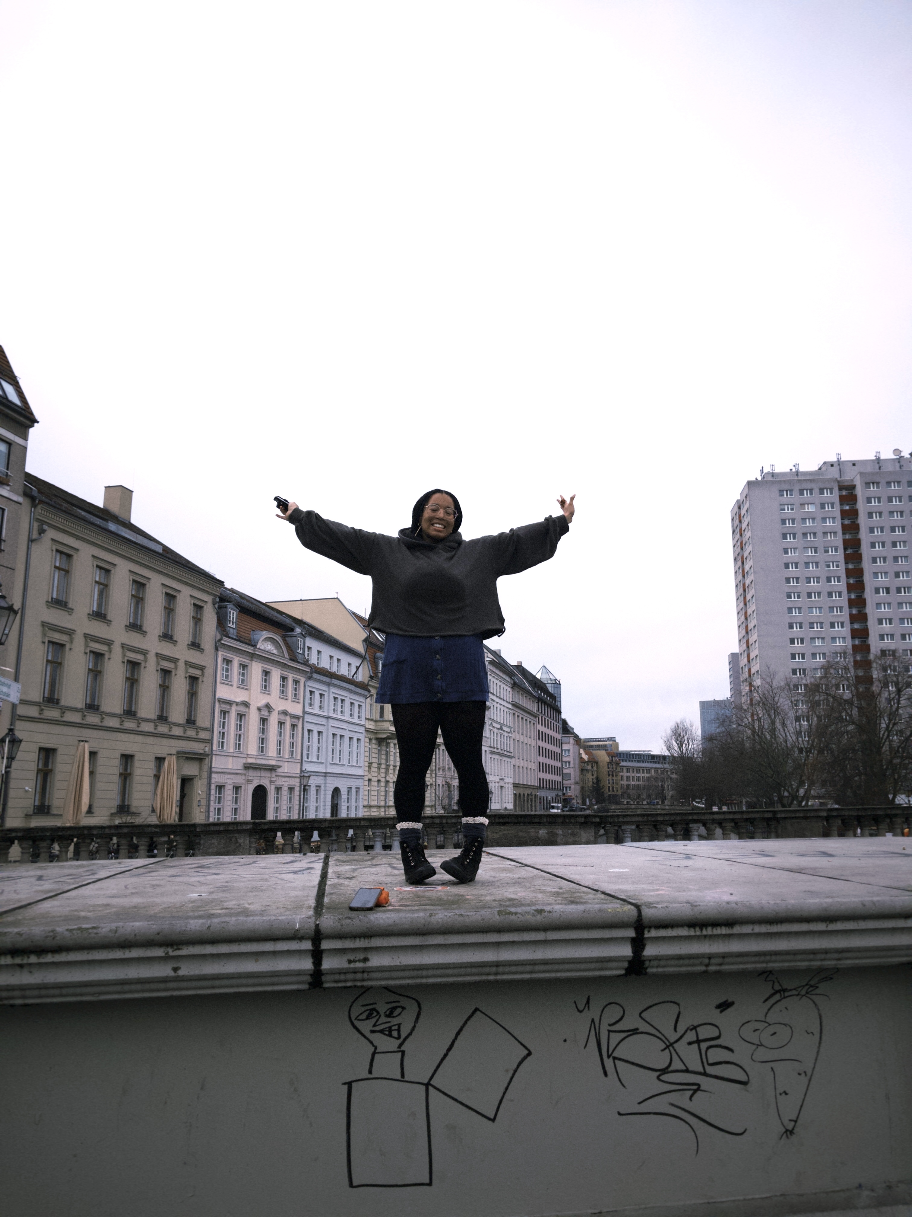 Lola stands on a short stone wall with street art on it. Her arms are spread wide and she's smiling.