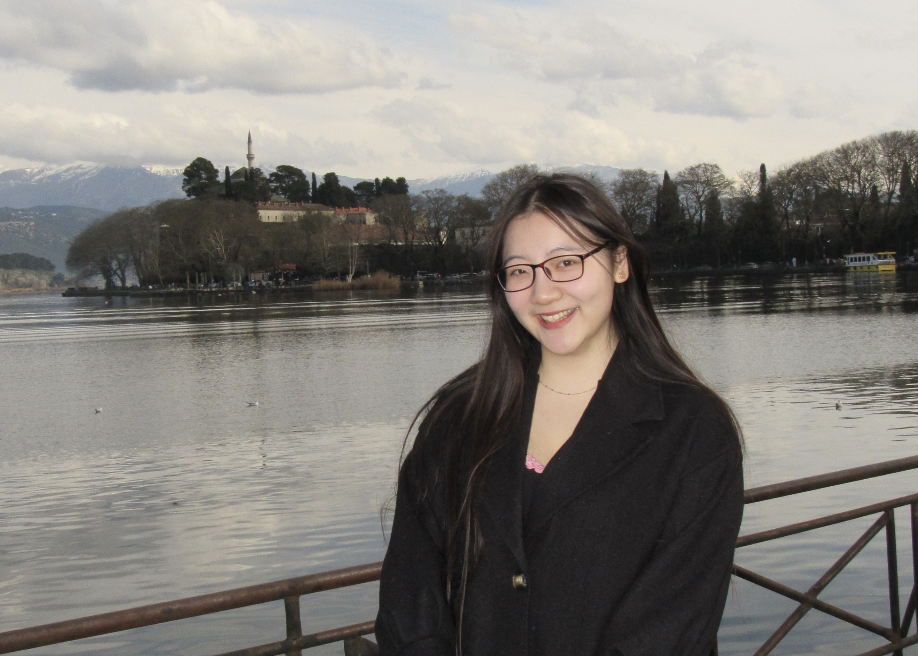 Michelle stands in front of a body of water with the cityscape of Thessaloniki behind her.