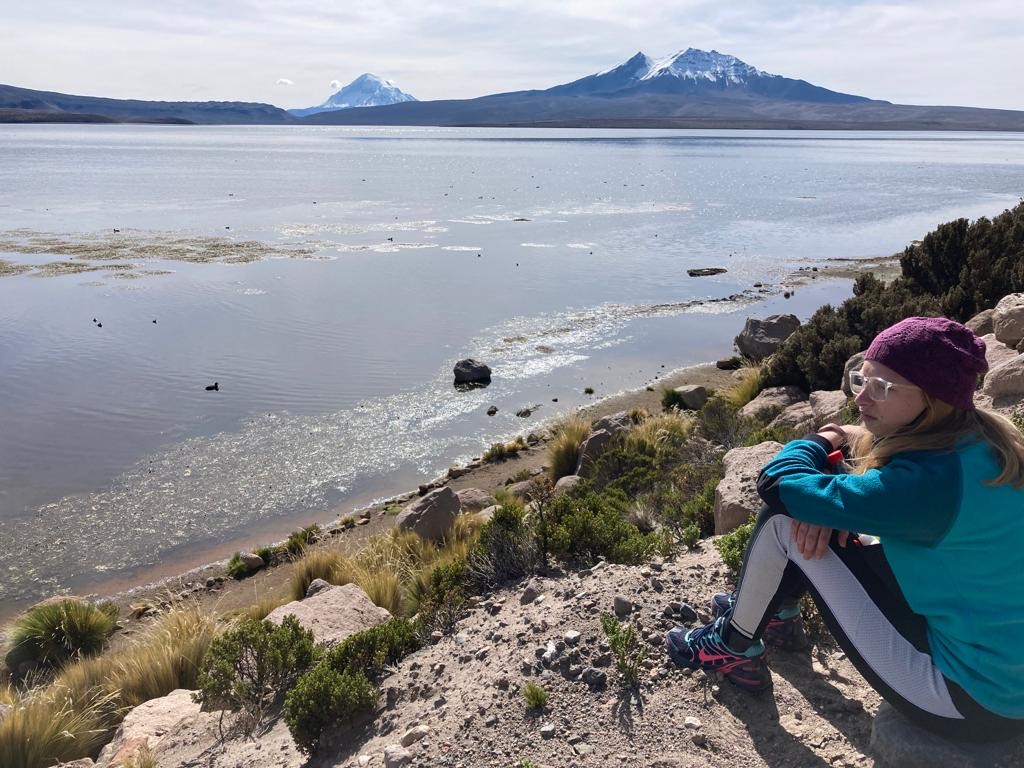 Rynn sits in profile on the right side of the image, looking out over water to mountains in the distance.