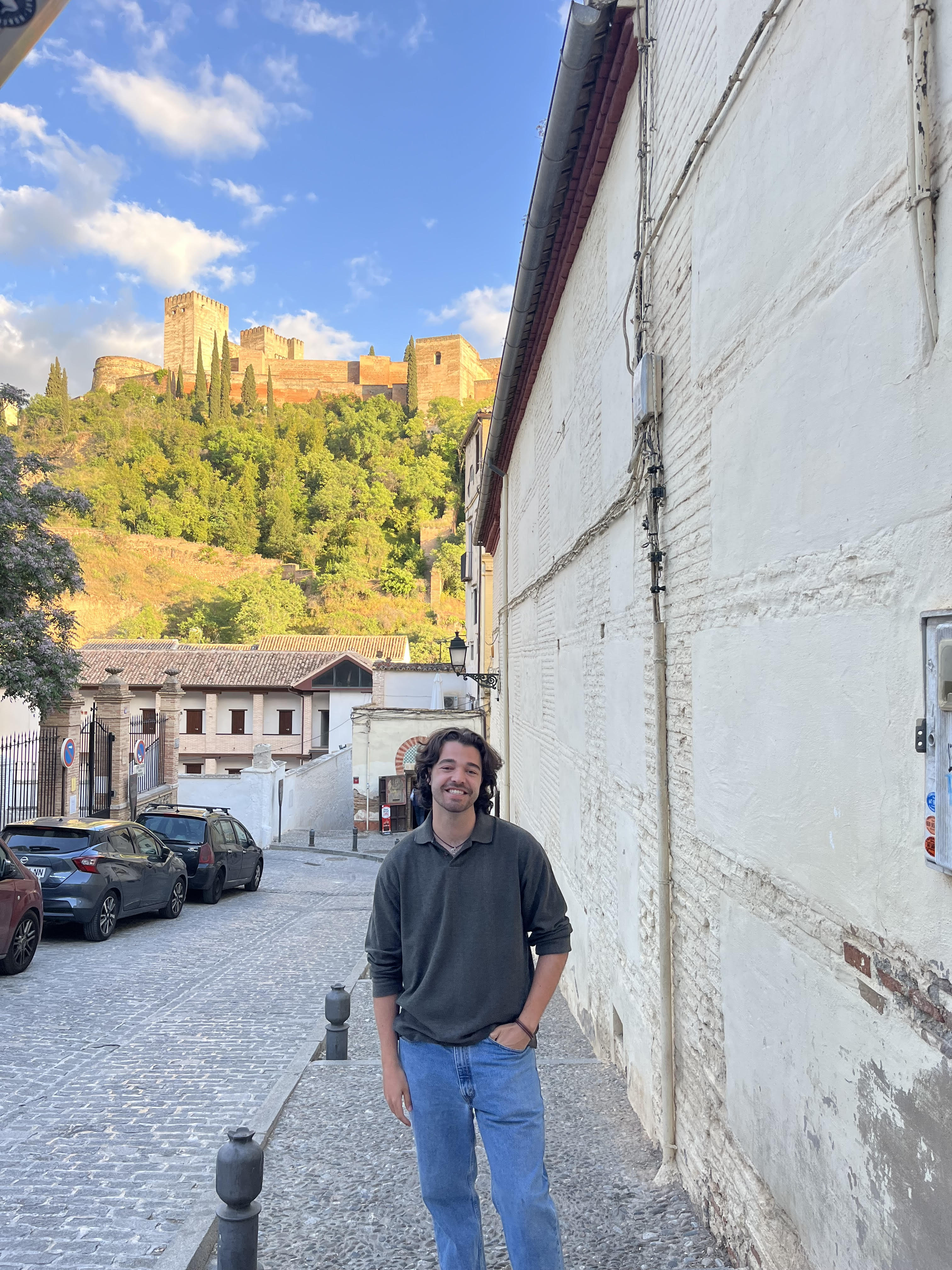 Spencer stands on a cobblestone street with the lush green trees and the Alhambra on a hill behind him.