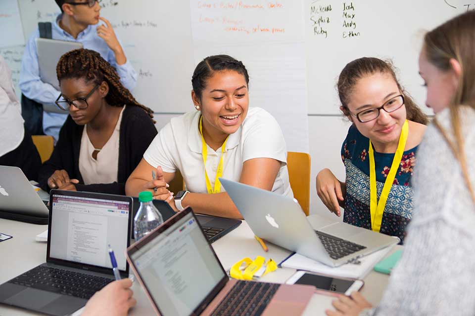 Students sit around a table in a classroom while a professor looks inquisitively at the whiteboard behind them
