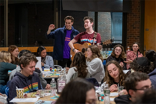 A student plays guitar and sings in front of a room of students seated at tables