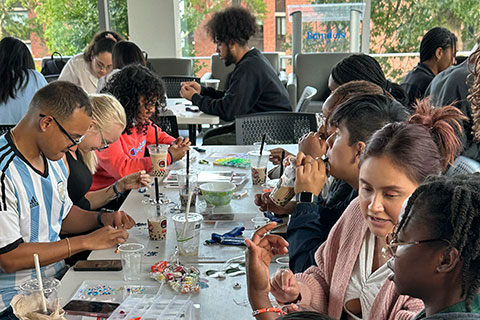 A group of people sit at a long table making bracelets and drinking boba tea.