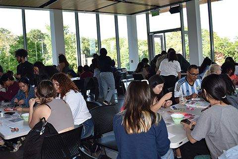 A group of people sit around various tables in a room talking and making bracelets.